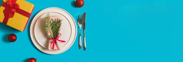 Banner with served plate and cutlery for celebration of Christmas and New Year. On plate is napkin with a Christmas tree branch, red balls. Flatlay banner on blue background with balls, gift box - Photo, Image