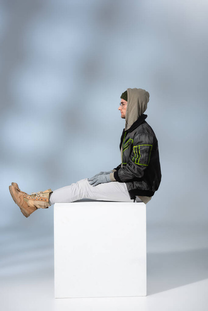 vista laterale dell'uomo di tendenza in cappello, guanti e anorak seduto su cubo bianco su grigio  - Foto, immagini