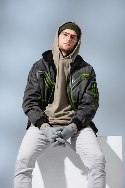 young trendy man in hat and anorak sitting on white cube with clenched hands in gloves on grey  - Фото, изображение