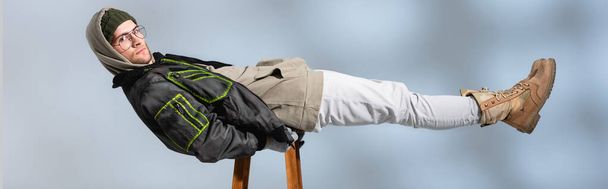 young trendy man in hat and anorak lying horizontally on wooden stool on grey, banner - Foto, Imagem