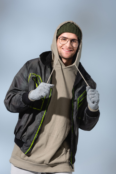 fashionable man in glasses, hat, anorak and gloves smiling on grey - Photo, Image