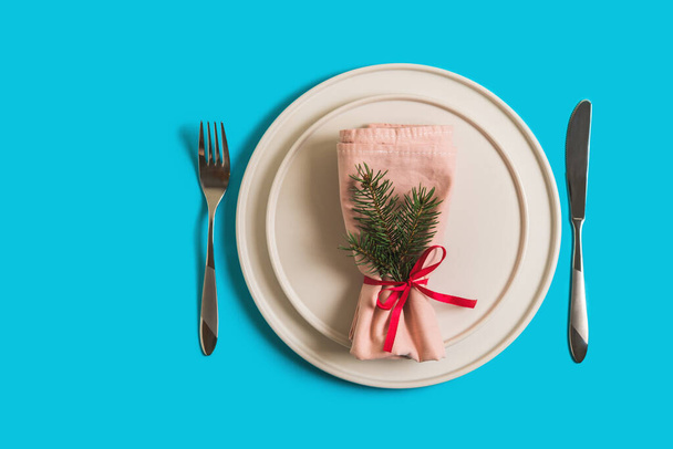 Served table with empty plate and cutlery for celebration of Christmas and New Year. On plate is napkin with Christmas tree branch. Flatlay on bright blue background. Top view - Photo, Image