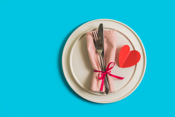 Served table with plate and cutlery for the celebration of Valentine's Day. On the plate is napkin with paper heart. Flatlay on bright blue background. Top view - Photo, Image