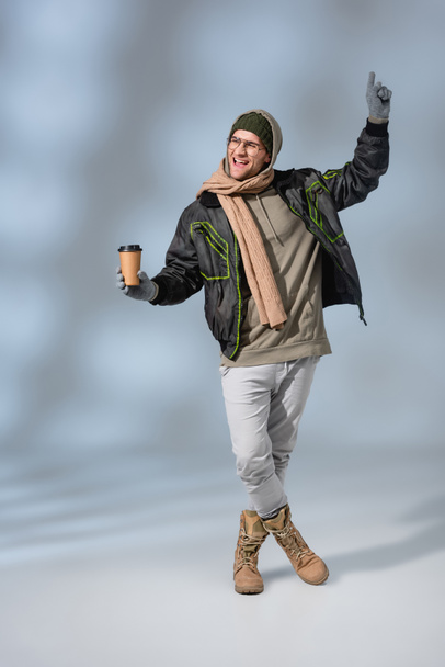 full length of excited man in hat, hoodie and anorak κρατώντας χάρτινο κύπελλο σε γκρι - Φωτογραφία, εικόνα