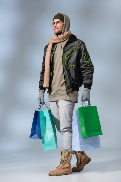 full length of smiling man in winter outfit holding shopping bags on grey - Photo, Image