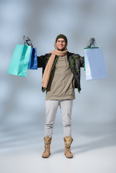 full length of happy man in parka holding shopping bags on grey - Zdjęcie, obraz