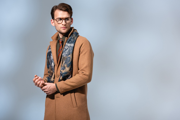 hombre de moda en gafas, bufanda y abrigo de invierno mirando hacia otro lado en gris - Foto, imagen