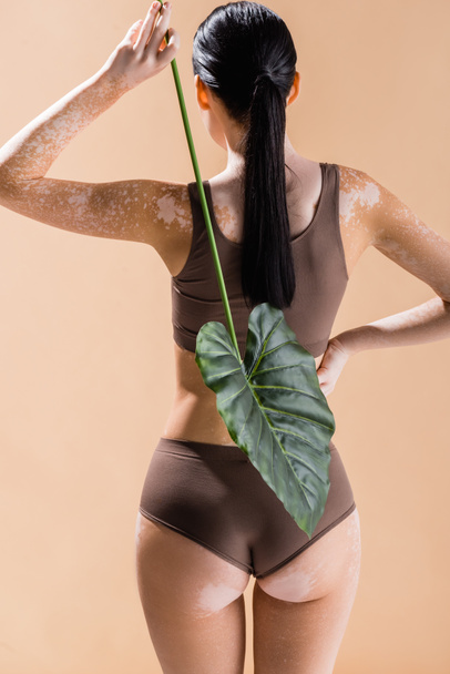 Rückseite der jungen schönen Frau mit Vitiligo posiert mit grünem Blatt isoliert auf beige - Foto, Bild