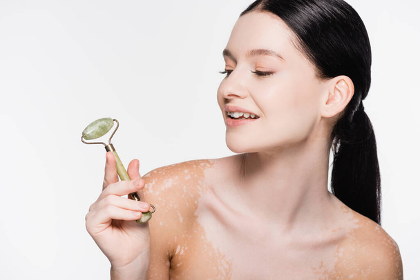 sorrindo jovem bela mulher com vitiligo segurando rolo de jade no rosto isolado no branco - Foto, Imagem