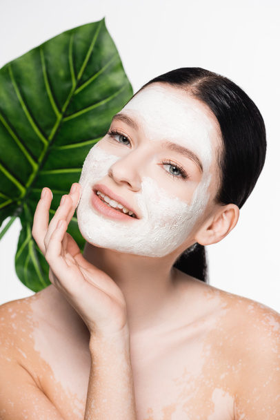 young beautiful woman with vitiligo and clay mask on face with blurred leaf on background isolated on white - Photo, Image