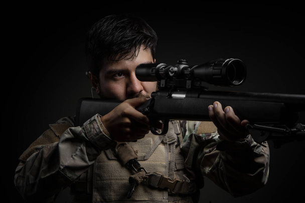Jongeman met een baard staand, vasthoudend en richtend met een geweer, gebruikt een oortelefoon en camouflage kleding, studio - Foto, afbeelding