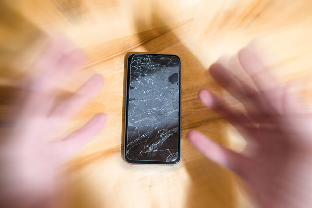 Concept of falling smart phone with broken screen. Top view on wooden desk background. Cracked, shattered lcd touch screen on modern cellphone. Gadget needs repairing - Photo, Image