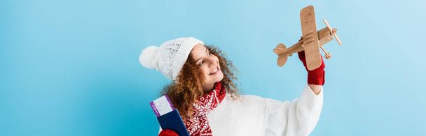 happy young woman in hat and sweater holding passport and toy plane on blue, banner - Fotografie, Obrázek