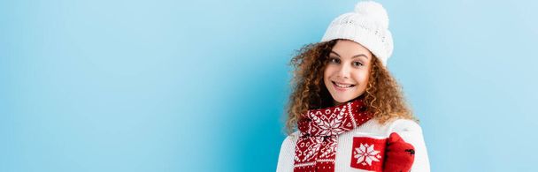 cheerful young woman in hat and sweater holding cup in knitted holder on blue, banner - Photo, Image