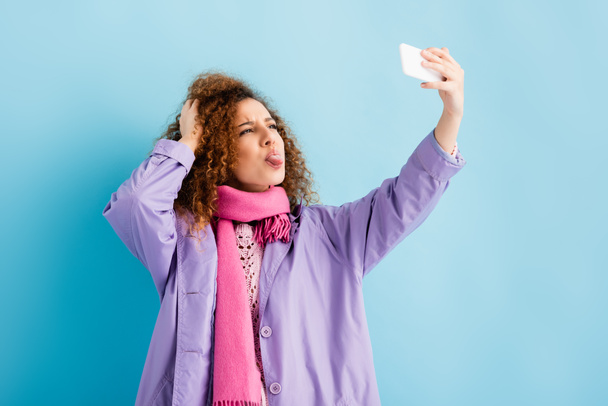 curly young woman in winter coat and pink knitted scarf holding smartphone and sticking out tongue while taking selfie on blue - Foto, Imagem