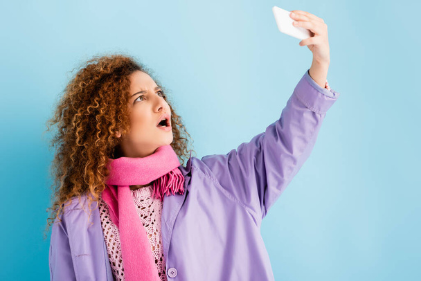 Schockierte junge Frau in Wintermantel und rosa Strickschal hält Smartphone in der Hand und macht Selfie auf blauem Grund - Foto, Bild