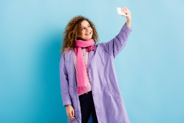 happy young woman in winter coat and pink knitted scarf holding smartphone and taking selfie on blue - Foto, Bild