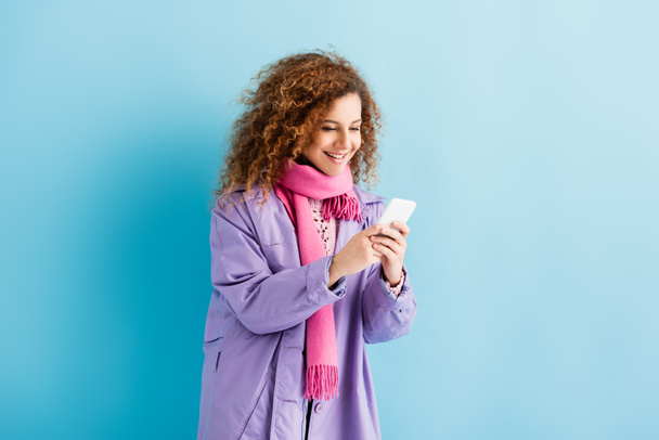 happy young woman in winter coat and pink knitted scarf texting on smartphone on blue - Photo, Image