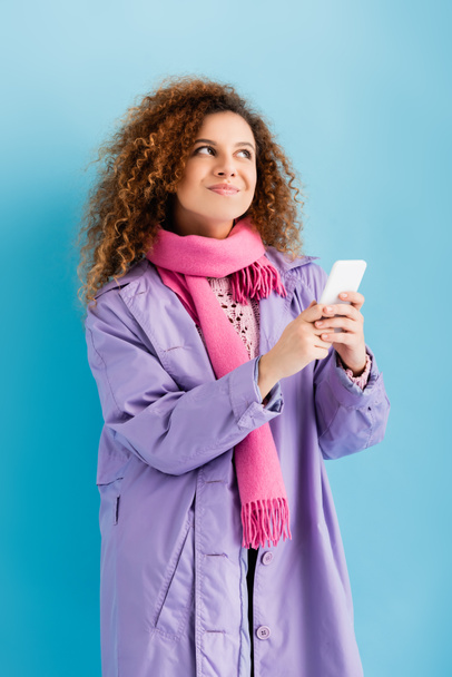 pensive young woman in winter coat and pink knitted scarf holding smartphone on blue - Foto, Imagen