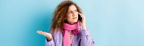 curly young woman in winter coat and pink knitted scarf talking on smartphone on blue, banner - Photo, Image