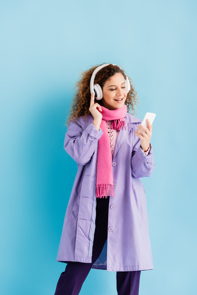 šťastná mladá žena v bezdrátových sluchátkách, zimní kabát a růžová pletená šála pomocí smartphonu na modré - Fotografie, Obrázek