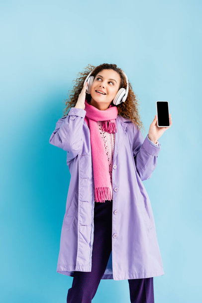 onnellinen nuori nainen langattomissa kuulokkeissa, talvitakki ja vaaleanpunainen neulottu huivi tilalla älypuhelin tyhjällä näytöllä sinisellä - Valokuva, kuva