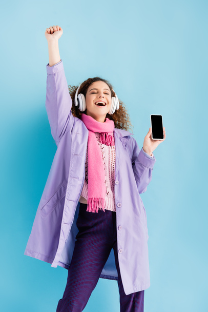 ενθουσιασμένοι νεαρή γυναίκα σε ασύρματα ακουστικά, χειμωνιάτικο παλτό και ροζ πλεκτό μαντήλι κρατώντας smartphone με λευκή οθόνη στο μπλε - Φωτογραφία, εικόνα