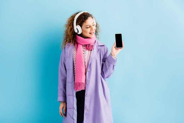 glückliche junge Frau in kabellosen Kopfhörern, Wintermantel und rosa Strickschal mit Blick auf Smartphone mit leerem Bildschirm auf blauem Hintergrund - Foto, Bild