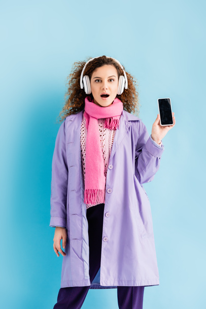 geschokt jonge vrouw in draadloze hoofdtelefoon, winterjas en roze gebreide sjaal holding smartphone met blanco scherm op blauw - Foto, afbeelding