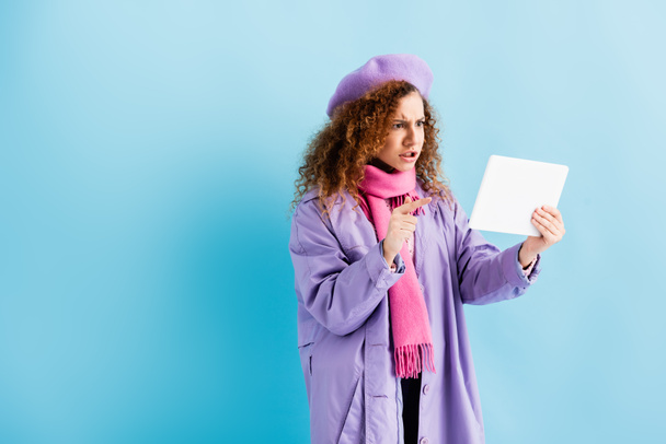 jeune femme insatisfaite en manteau d'hiver, béret et écharpe tricotée rose pointant du doigt la tablette numérique sur bleu - Photo, image