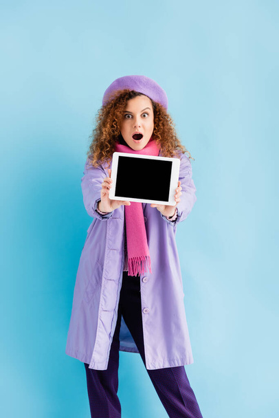 šokovaná mladá žena v zimním kabátě, baretu a růžovém pleteném šátku držící digitální tablet s prázdnou obrazovkou na modré - Fotografie, Obrázek