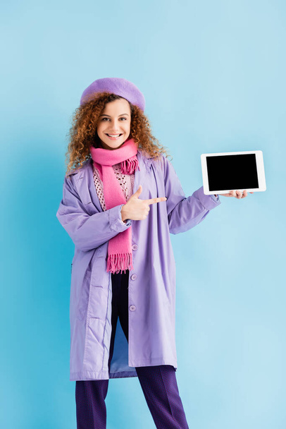 šťastná žena v zimním kabátě, baretu a růžové pletené šále ukazující prstem na digitální tabletu s prázdnou obrazovkou na modré - Fotografie, Obrázek