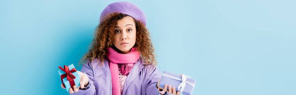 curly woman in winter coat, beret and pink knitted scarf holding christmas presents on blue, banner - Foto, afbeelding