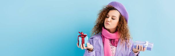 curly woman in winter coat, beret and pink knitted scarf choosing between christmas presents on blue, banner - Zdjęcie, obraz