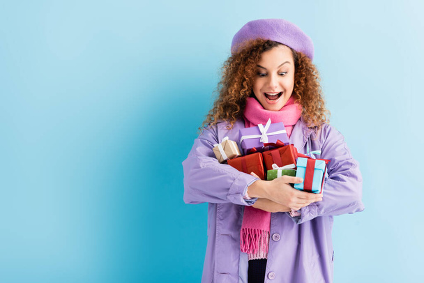 excited woman in winter coat, beret and pink knitted scarf holding christmas presents on blue  - Foto, Imagen