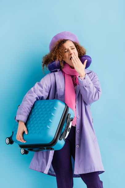 tired woman in beret, scarf, travel pillow and coat holding suitcase and yawning on blue - Fotoğraf, Görsel