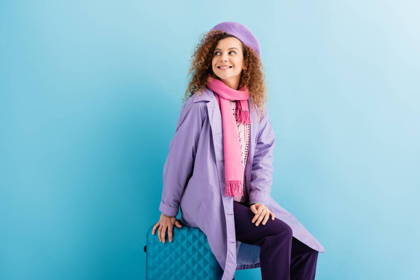 cheerful woman in beret, scarf, travel pillow and coat sitting on luggage on blue - Photo, Image