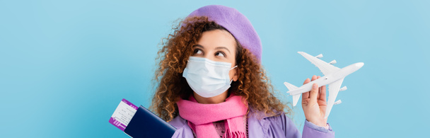donna riccia in berretto, sciarpa e maschera medica in possesso di passaporto e aereo giocattolo su blu, banner - Foto, immagini