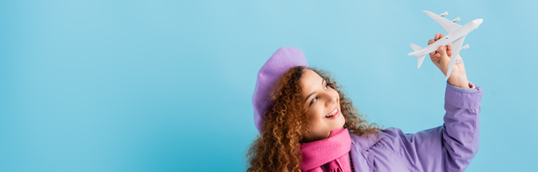 positive woman in beret, scarf and coat holding toy plane on blue, banner - Fotó, kép