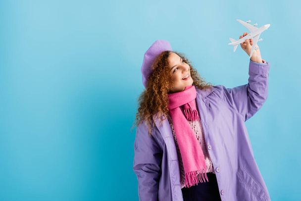 cheerful woman in beret, scarf and coat looking at toy plane on blue - Φωτογραφία, εικόνα