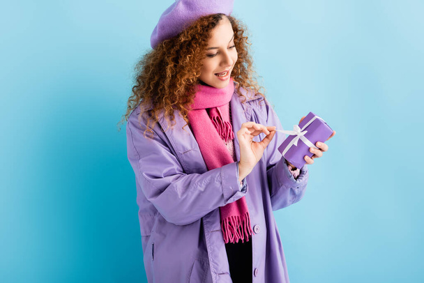happy woman in winter coat, beret and pink knitted scarf untying ribbon on christmas present on blue  - Foto, Imagen