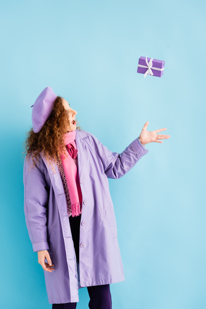 glückliche Frau in Wintermantel, Baskenmütze und rosa Strickschal wirft Weihnachtsgeschenk auf blau  - Foto, Bild