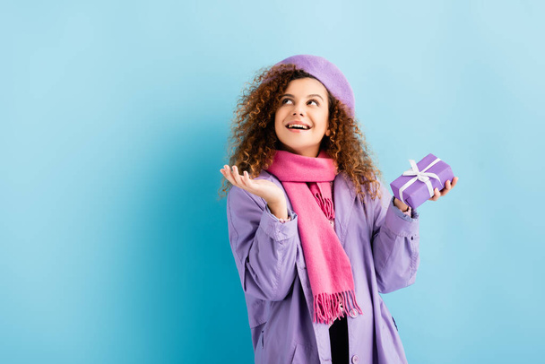 smiling woman in winter coat, beret and pink knitted scarf holding christmas present on blue  - Foto, Imagem