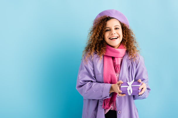 sorrindo jovem mulher no casaco de inverno, boina e lenço de malha rosa segurando Natal presente no azul  - Foto, Imagem