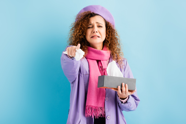 upset young woman in winter coat, beret and pink knitted scarf holding tissue box and napkin on blue  - Foto, immagini