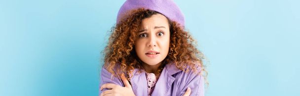 curly woman in beret freezing and embracing herself on blue background, banner - Photo, Image