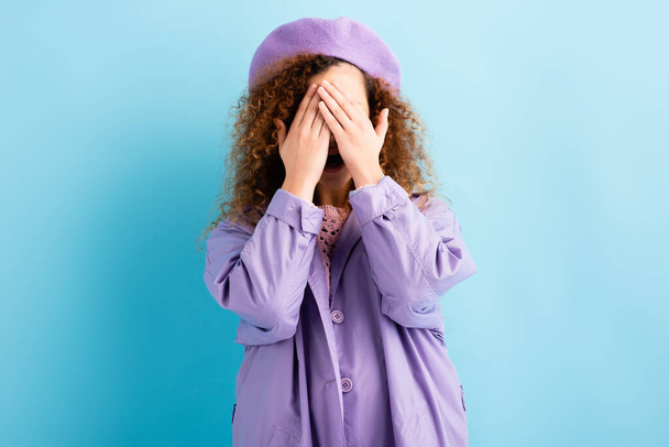 scared and curly woman in beret covering face with hands on blue - Фото, зображення