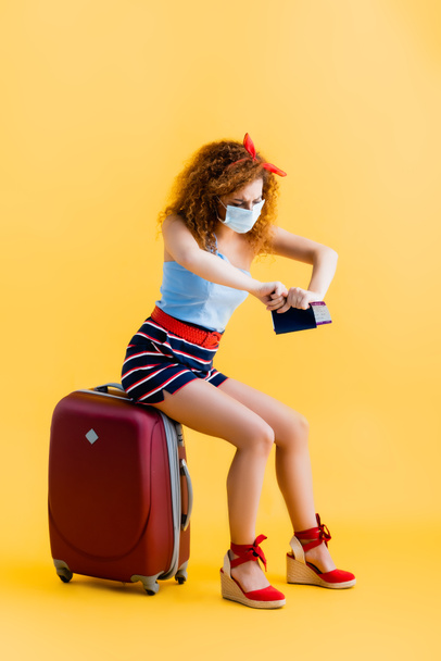 lockige junge Frau in medizinischer Maske zerreißt Pass, während sie auf Koffer auf gelb sitzt  - Foto, Bild