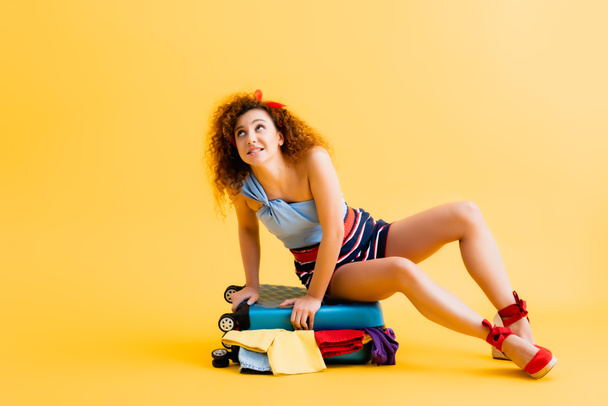 curly young woman sitting on suitcase with clothing on yellow - Photo, Image