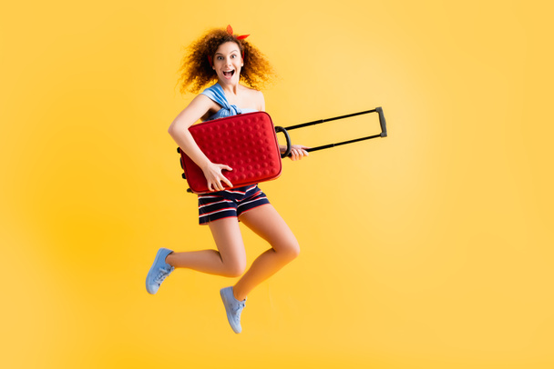 full length of happy woman in summer outfit and sneakers jumping with suitcase on yellow   - Photo, image